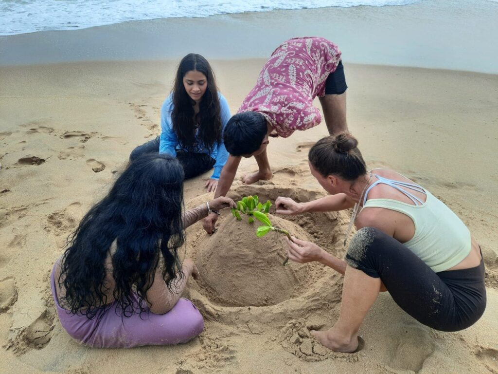 A small group kneeling together on a beach, shaping a mound of sand and planting a green sprig in it